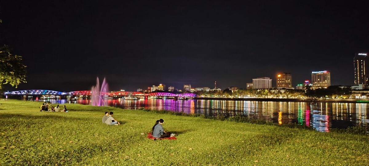 Calm river landscape along the Perfume River at night with people resting by the grassy riverbank in Hue, Vietnam
