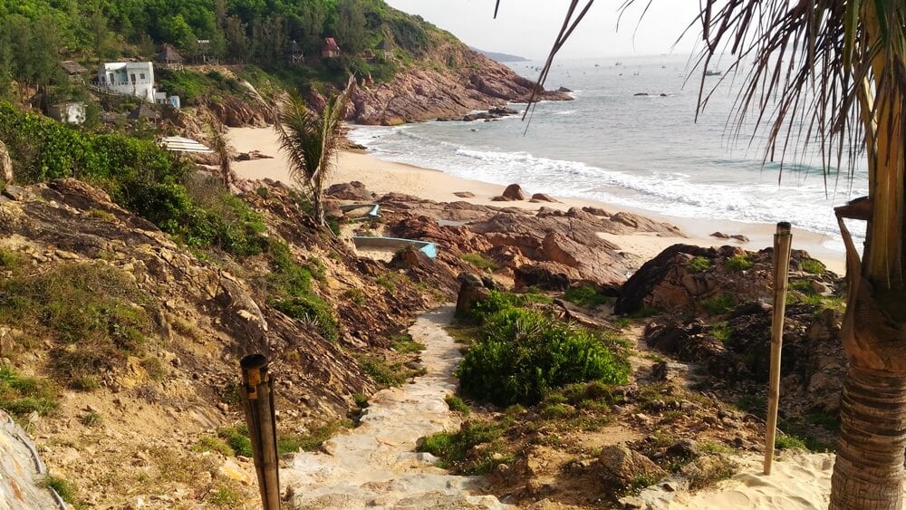 Walkway from a beachfront resort to the sea in Quy Nhon, with rocky shoreline, mountains, and coastal views